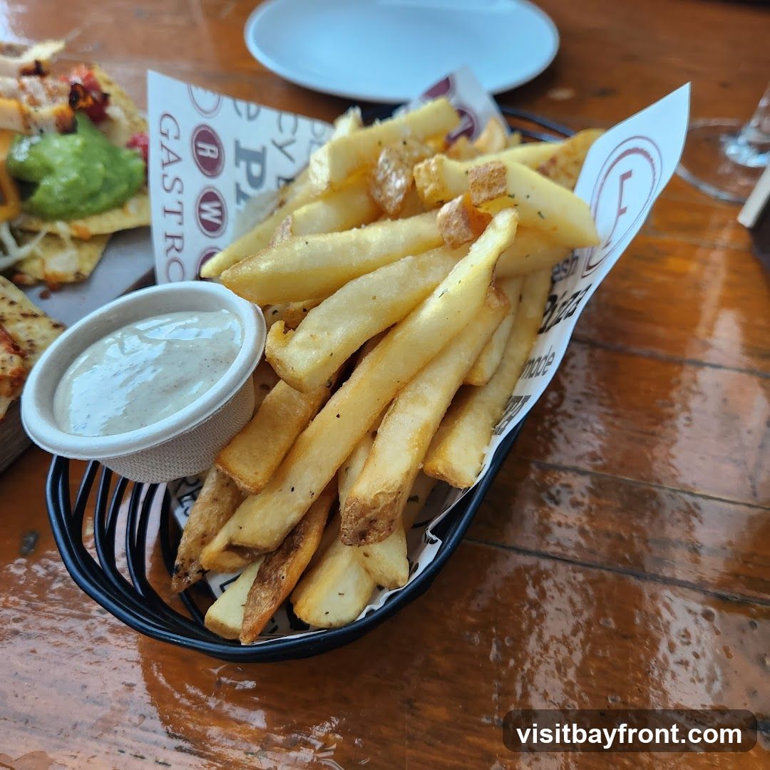 A black wire basket filled with golden-brown french fries sits on a wooden table. A small paper cup of creamy white dip is nestled among the fries. In the background, a plate with what appears to be nachos or tacos with guacamole is visible. The paper lining the basket has a repeating logo and text that includes "GASTRO" and "Pizza".