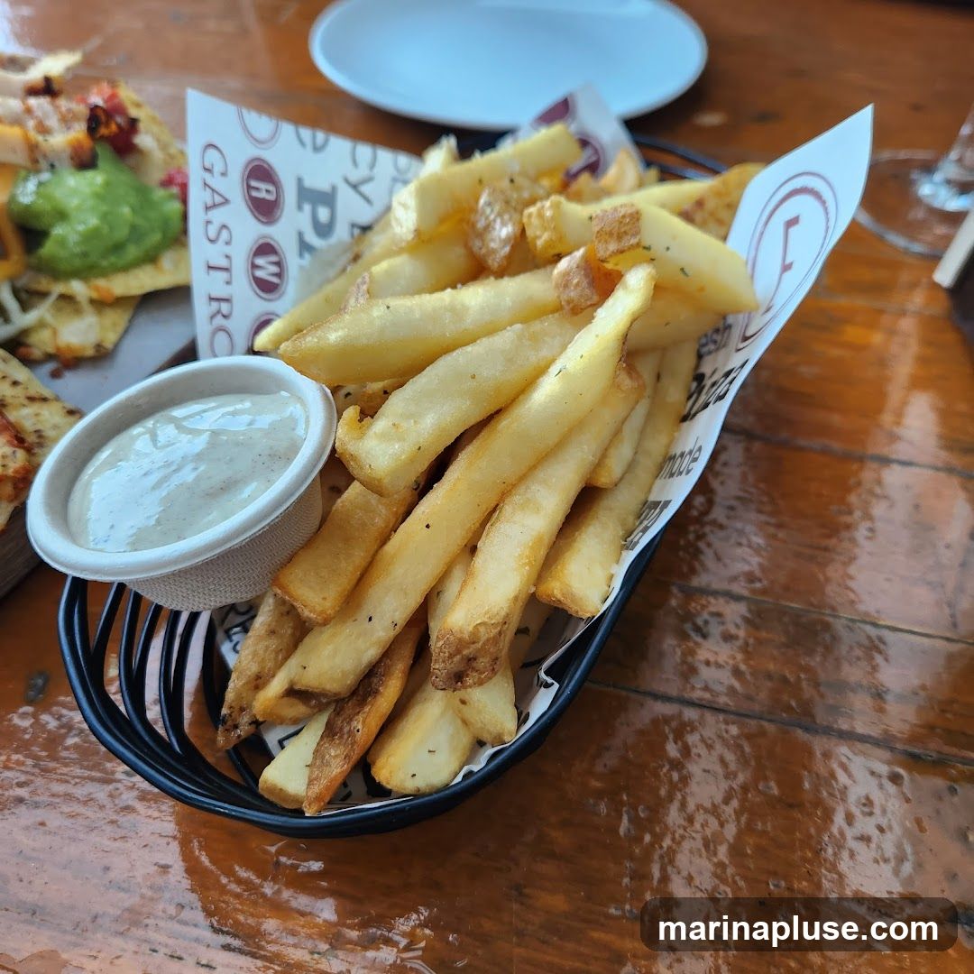 A black wire basket filled with golden-brown french fries sits on a wooden table. A small paper cup of creamy white dip is nestled among the fries. In the background, a plate with what appears to be nachos or tacos with guacamole is visible. The paper lining the basket has a repeating logo and text that includes "GASTRO" and "Pizza".