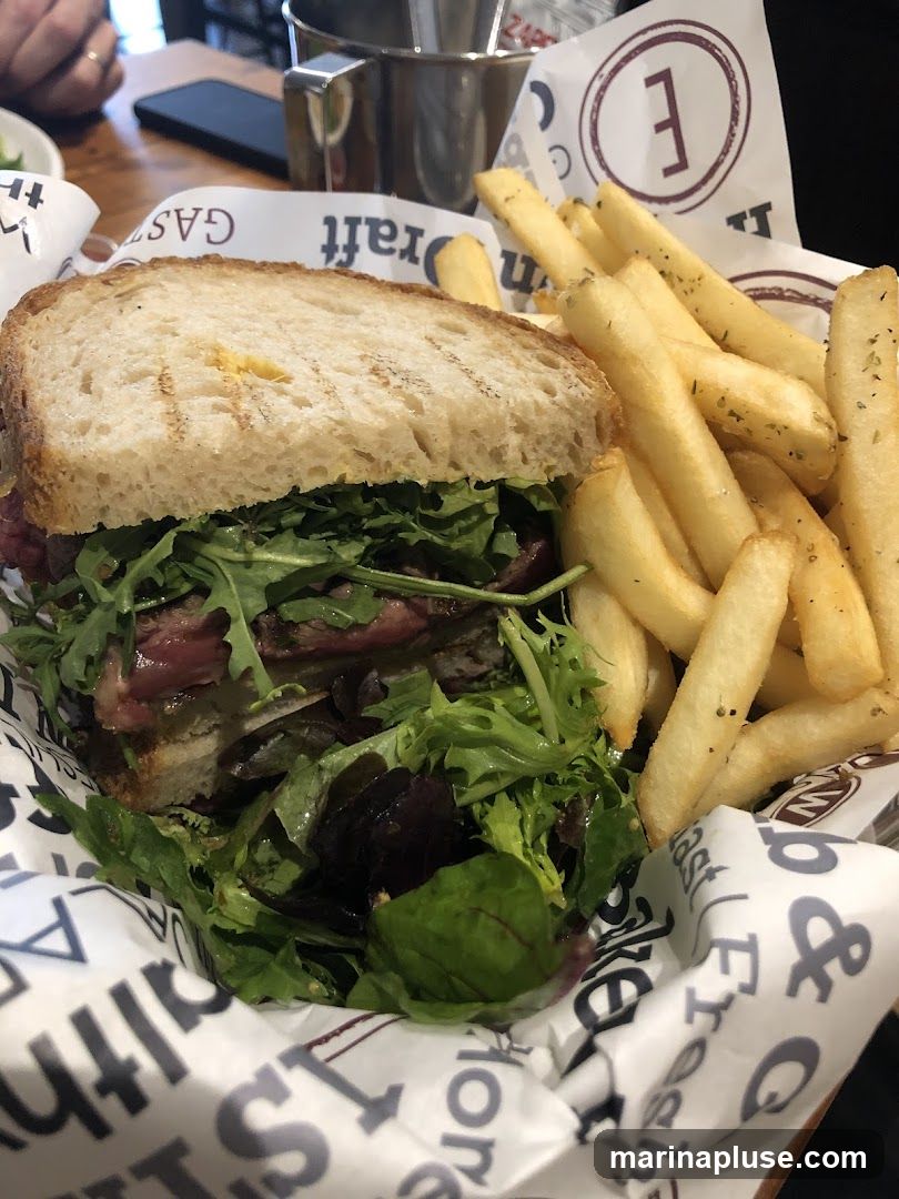 A close-up shot of a sandwich and french fries served in a basket lined with branded paper. The sandwich appears to be made with grilled bread, filled with sliced meat, and topped with fresh greens. The fries are golden brown and seasoned. The paper lining has text and a logo with the letter 'E' inside a circle.