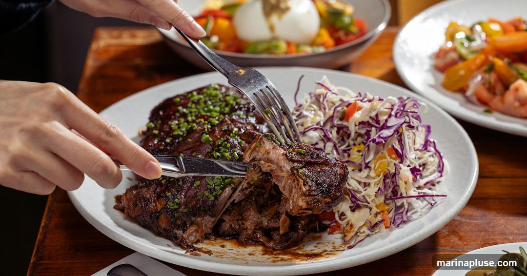 A close-up shot of a person cutting into a plate of braised ribs, garnished with chopped green herbs. The ribs are served with a side of coleslaw, which includes red cabbage, shredded white cabbage, and diced bell peppers. In the background, out of focus, are two other plates of food: one with a burrata cheese and tomato salad, and another with sliced tomatoes. The meal is presented on a wooden table.