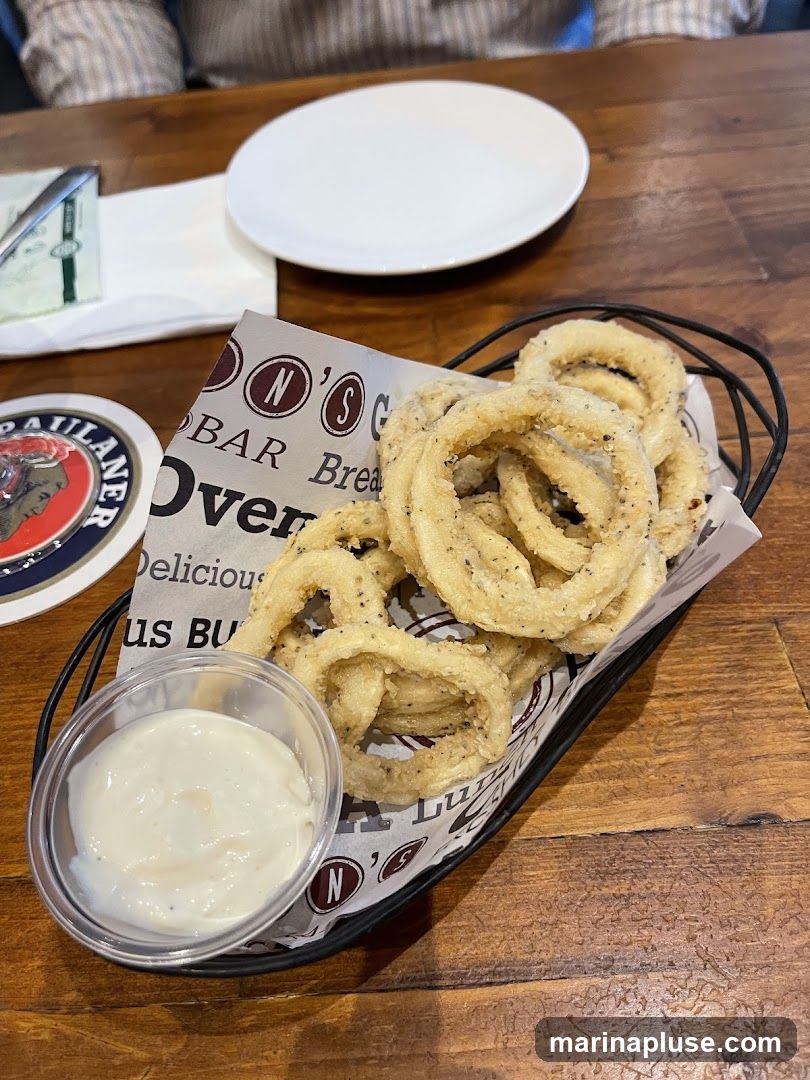 A basket of fried calamari rings served with a small dish of dipping sauce. The calamari is seasoned with black pepper. The basket is lined with paper that has a repeating logo with the letters 'N' and 'S' inside circles, and the words 'BAR' and 'Over'. A coaster with a logo featuring a man's profile and the word 'PAULANER' is visible to the left. A white plate and cutlery are on a wooden table.