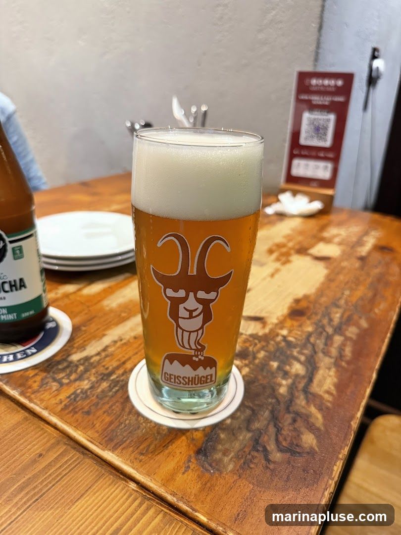 A pint glass filled with beer and a frothy head sits on a wooden table. The glass features a cartoonish goat logo with the text "GEISSHÜGEL" below it. To the left, a bottle of kombucha and a stack of white plates are visible. In the background, a QR code menu is displayed on a stand.
