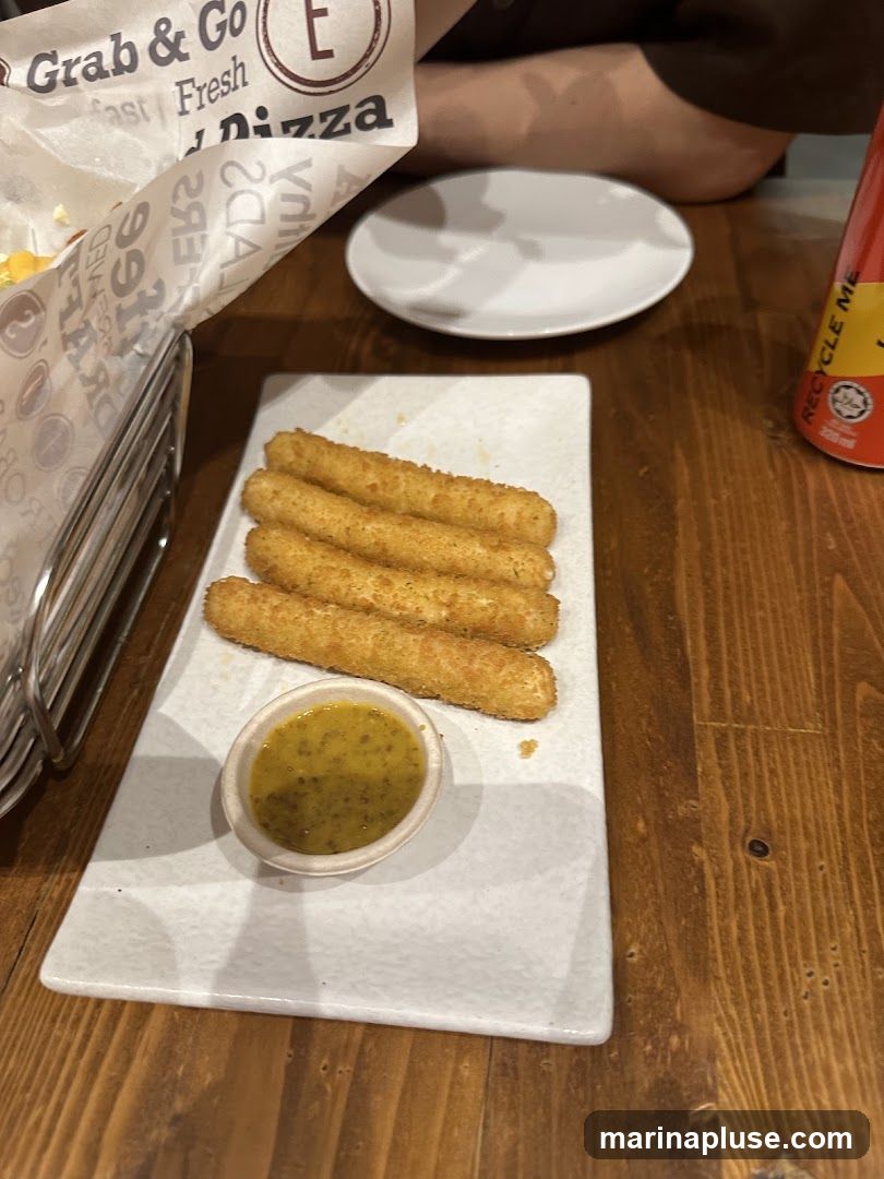 A plate of golden-brown mozzarella sticks served with a small bowl of dipping sauce sits on a wooden table. A white plate and a red can are also visible. A paper liner with text that includes "Grab & Go" and "Pizza" is in the background.