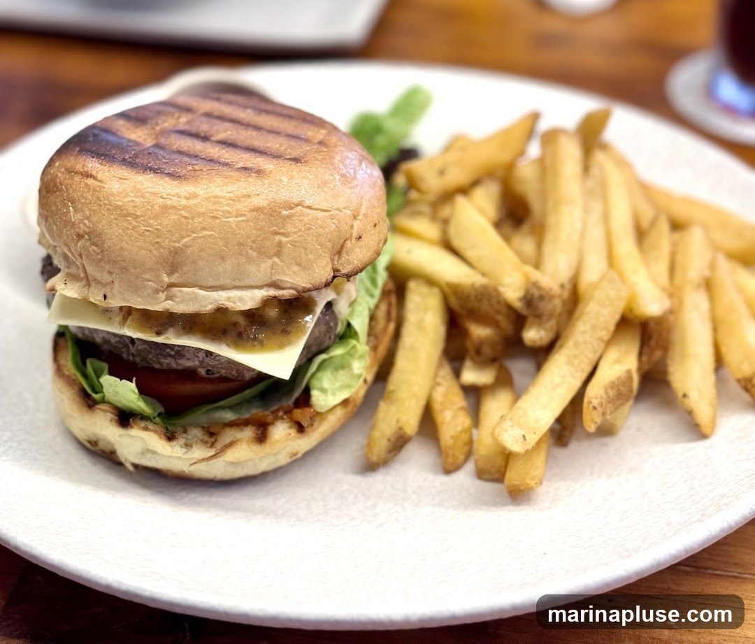 A close-up shot of a gourmet burger served with a side of golden-brown french fries on a white plate. The burger features a toasted bun, a thick beef patty, melted cheese, fresh lettuce, and tomato, topped with a savory sauce. The fries are thick-cut and appear seasoned.