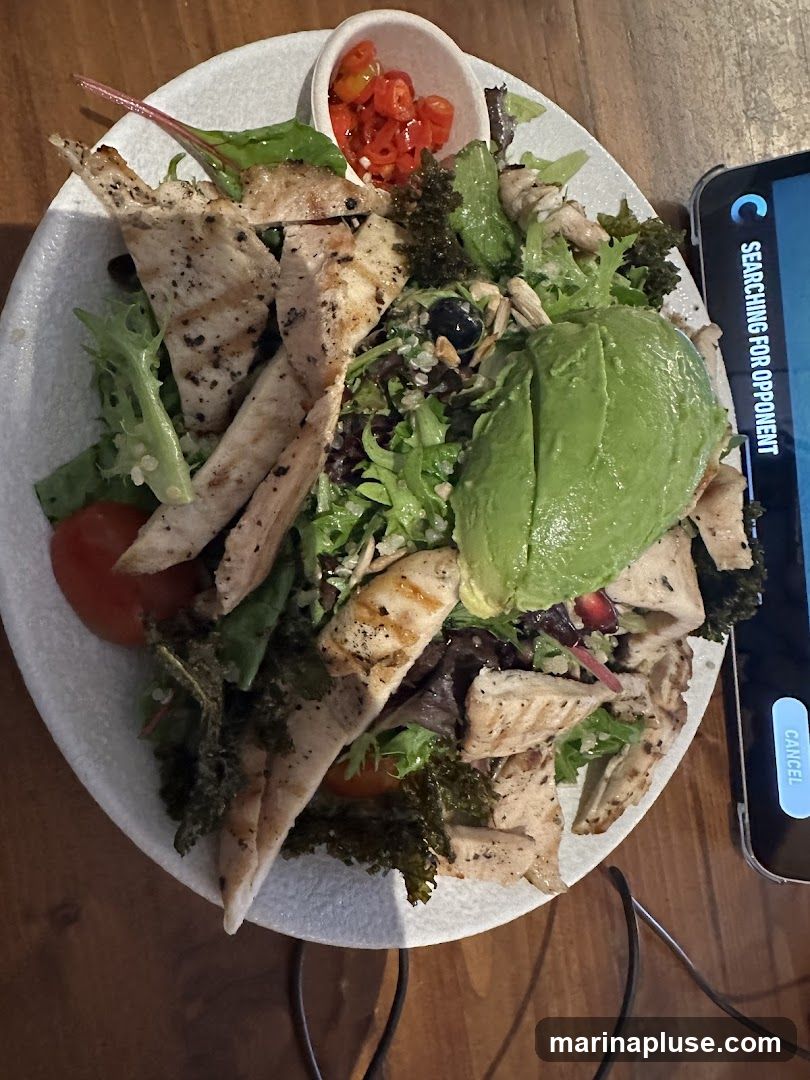 A close-up overhead shot of a chicken salad served on a white plate. The salad contains grilled chicken strips, mixed greens, avocado slices, tomatoes, and chopped red chilies in a small side dish. A portion of a smartphone screen is visible on the right side of the image.