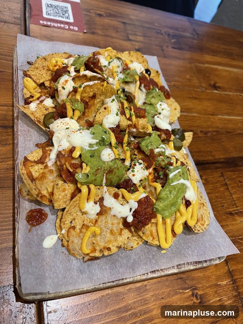 A close-up shot of a large serving of nachos on a wooden table. The nachos are topped with melted cheese, guacamole, salsa, sour cream, and chives. A small sign with a QR code and the text "Go Cashless" is visible in the upper left corner.
