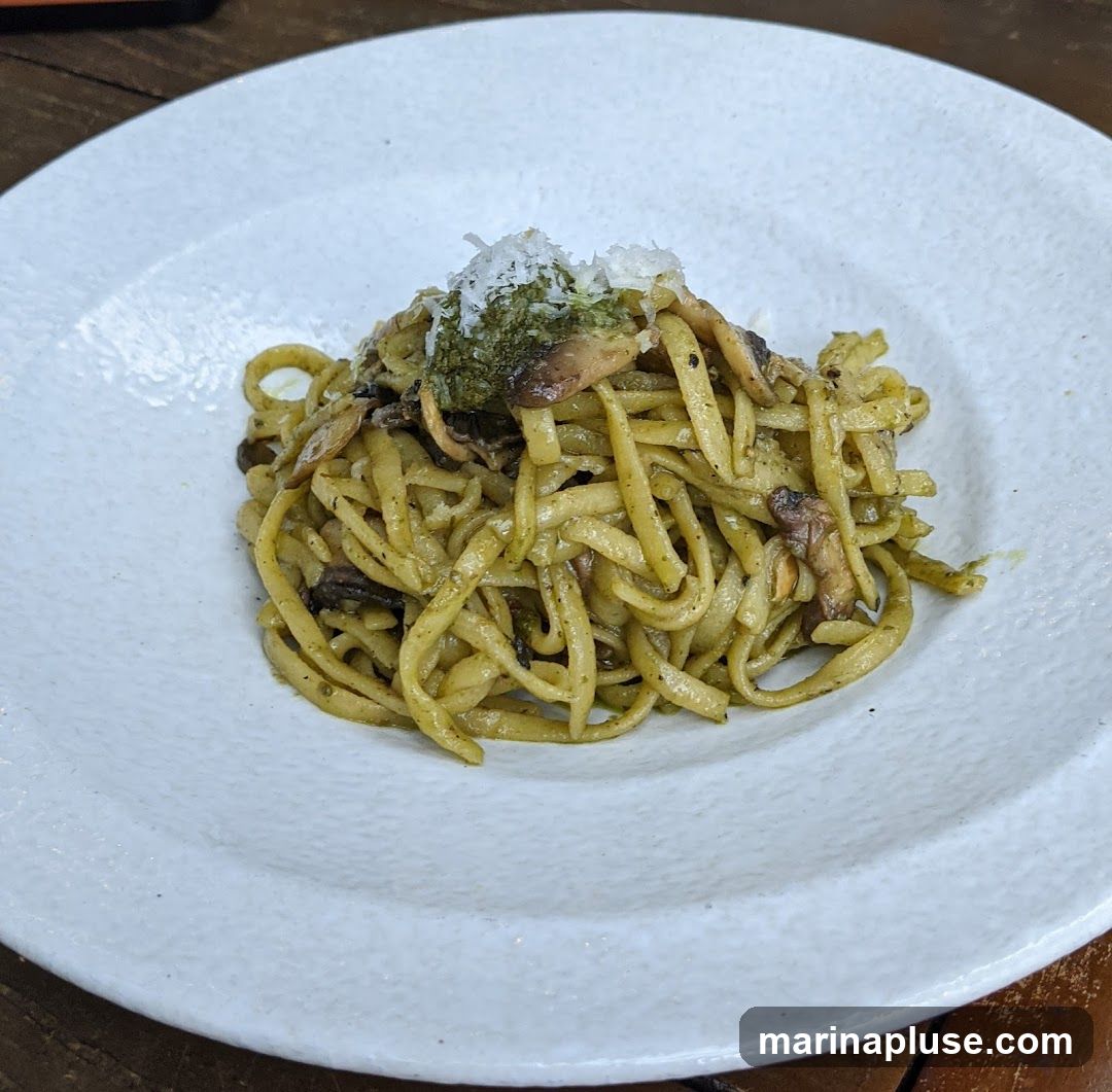 A close-up shot of a plate of pasta with mushrooms and pesto sauce. The pasta appears to be linguine or fettuccine, coated in a vibrant green pesto. Sliced mushrooms are mixed throughout the pasta, and the dish is topped with a sprinkle of grated cheese and a dollop of pesto. The plate is white and textured, and it sits on a wooden surface.