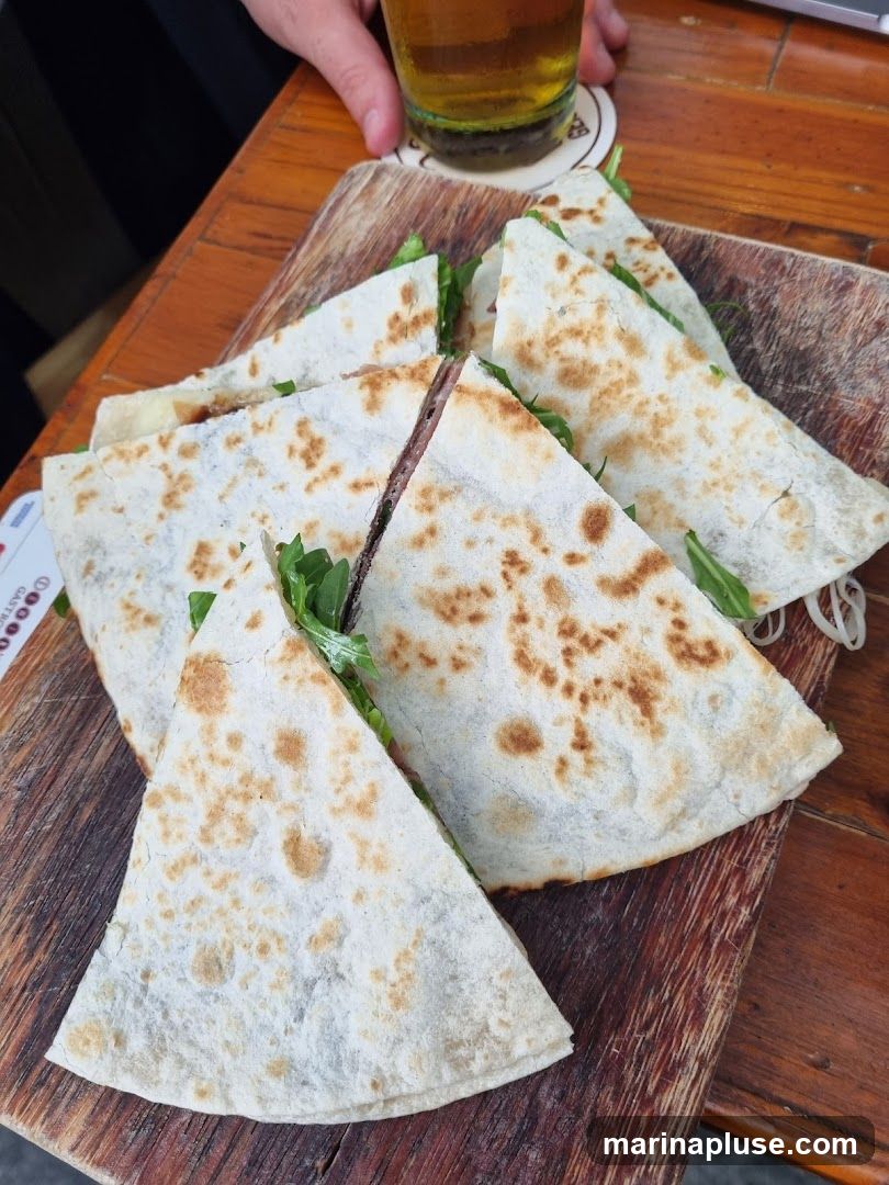 A wooden board with several triangular quesadillas filled with greens and meat. A glass of beer and a hand are visible in the background.