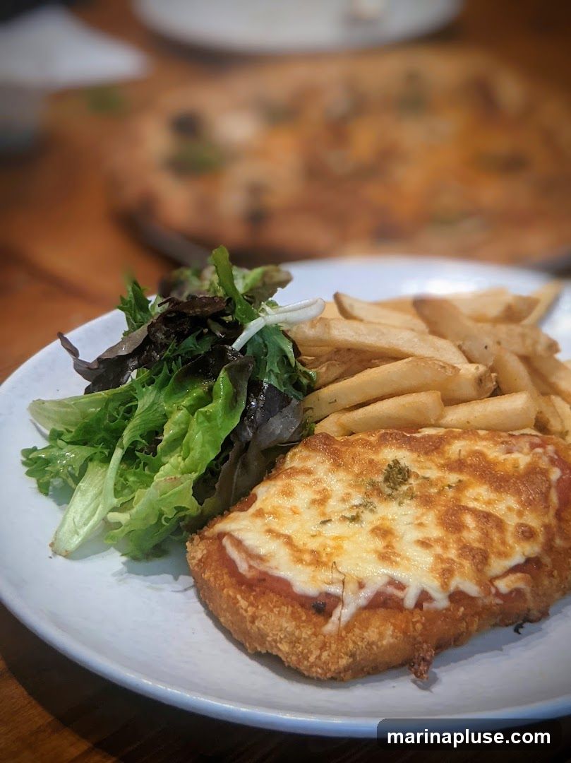 A close-up shot of a chicken parmigiana served with a side of french fries and a small green salad. The chicken is breaded and topped with tomato sauce and melted cheese. In the background, a pizza is out of focus.