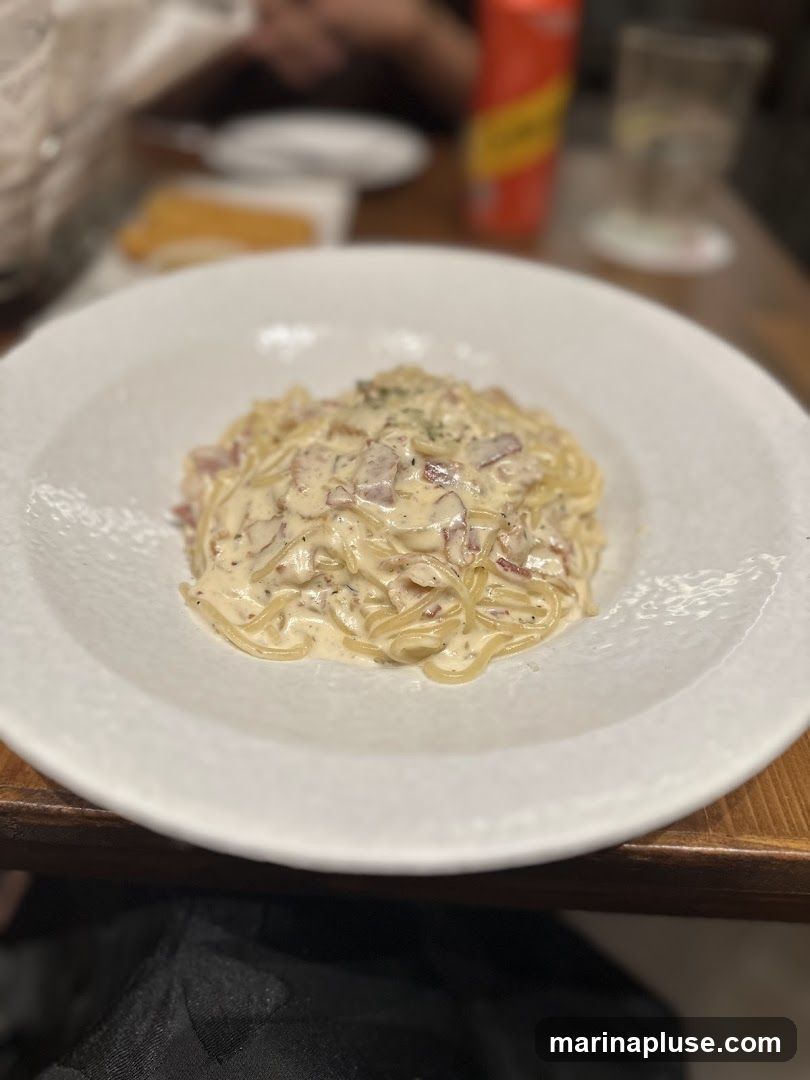 A close-up shot of a white bowl filled with spaghetti carbonara. The pasta is coated in a creamy sauce with visible pieces of bacon or pancetta and sprinkled with herbs. In the blurred background, a red and yellow can, a glass, and other diners are visible.