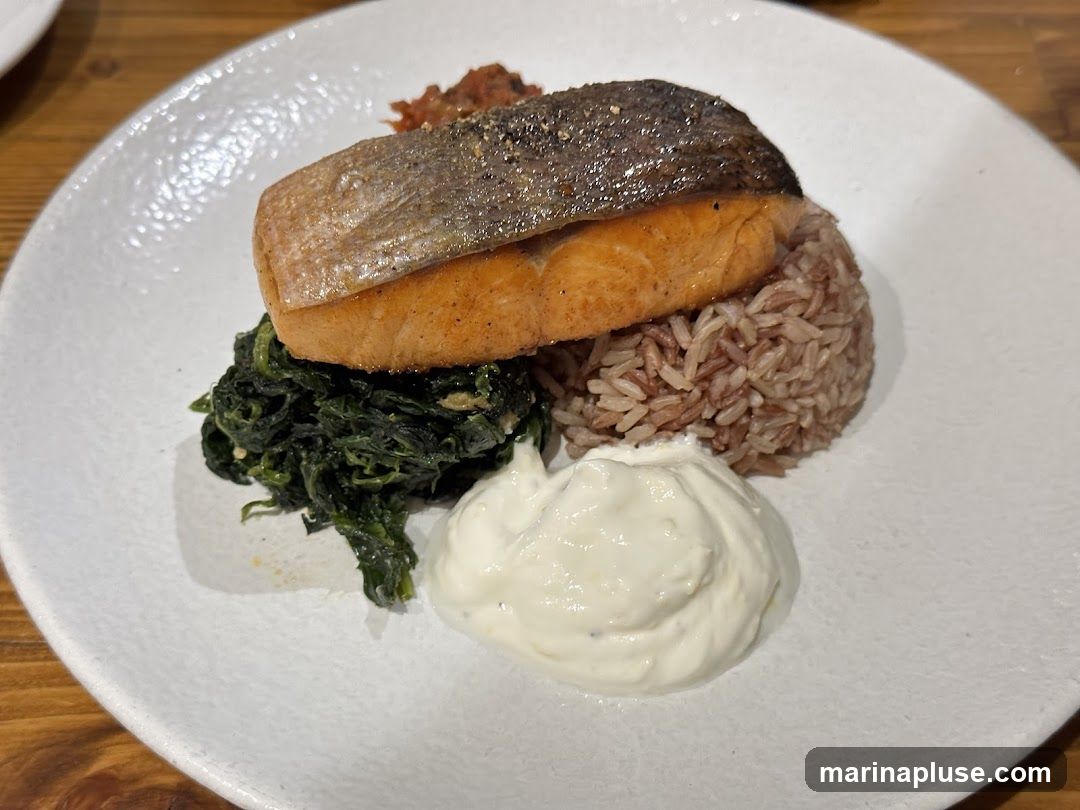 A plate of food featuring a piece of salmon with crispy skin, served with a side of brown rice, sautéed spinach, a dollop of white sauce, and a small portion of red relish.