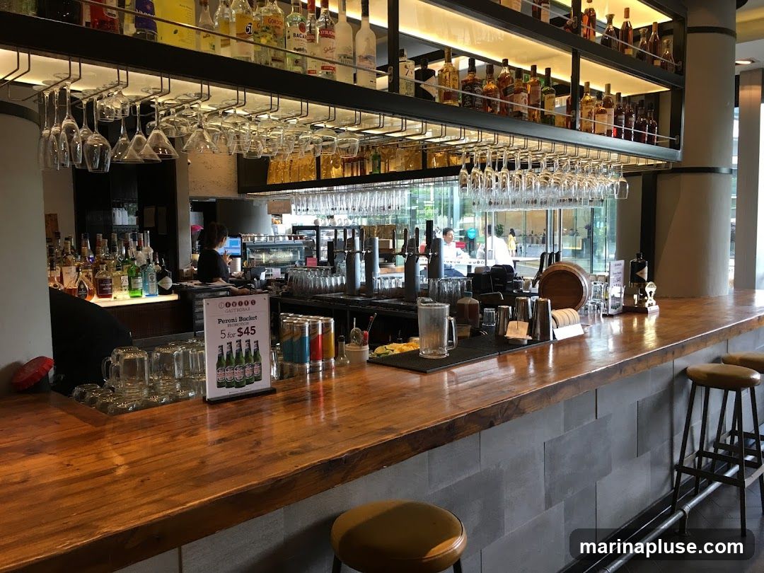 A well-lit bar interior featuring a long wooden counter with various bottles of alcohol displayed on shelves above. Numerous wine glasses hang from racks overhead. Behind the counter, a barista is visible working at a coffee machine. Several bar stools are positioned along the counter, and a promotional sign for a "Peroni Bucket" is displayed. The overall ambiance suggests a modern and inviting establishment.