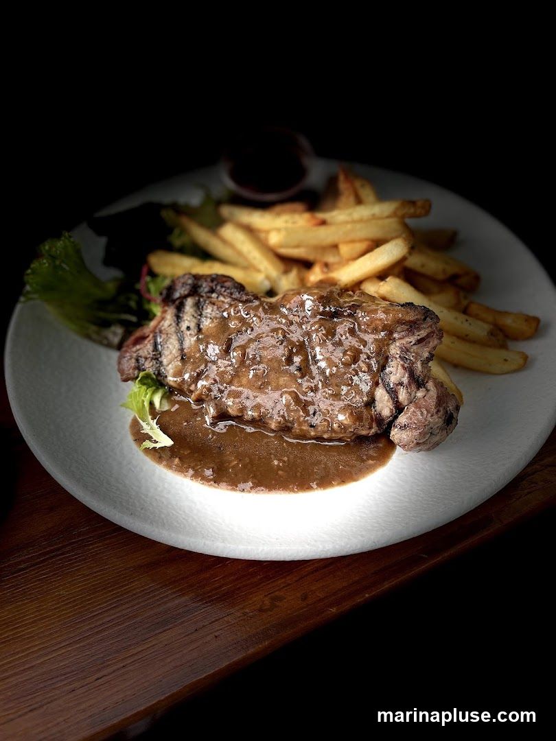 A plate of steak and fries is presented on a wooden table. The steak is covered in a rich brown sauce and served with a side of golden french fries and a small salad. A small dish of sauce is visible in the background.
