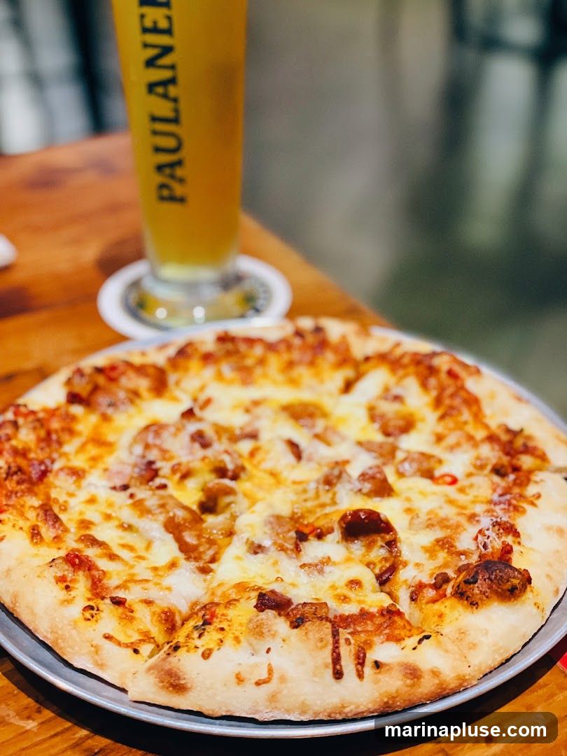 A close-up shot of a pizza with melted cheese and toppings, served on a metal plate. A tall glass of beer with the Paulaner logo is visible in the background, placed on a coaster. The food is on a wooden table.