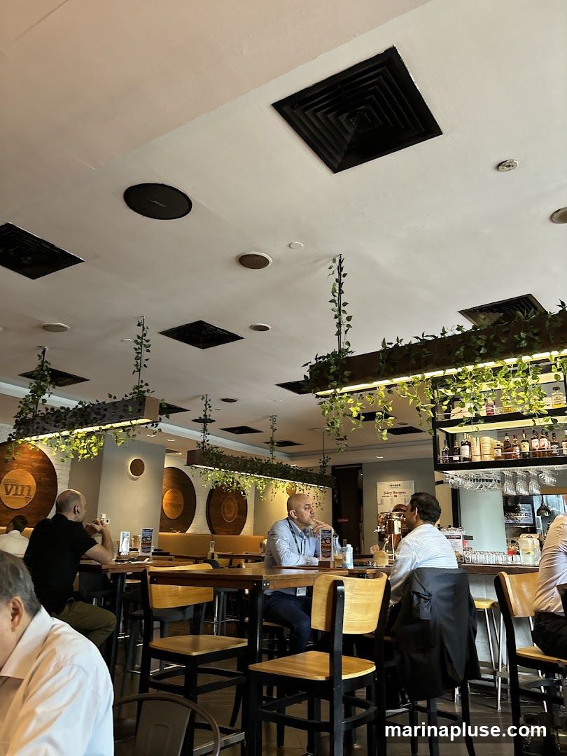 The image shows the interior of a restaurant with several patrons seated at tables. The decor includes wooden accents, hanging plants, and a bar area stocked with bottles and glassware. A circular logo with the word "vin" is visible on the wall.