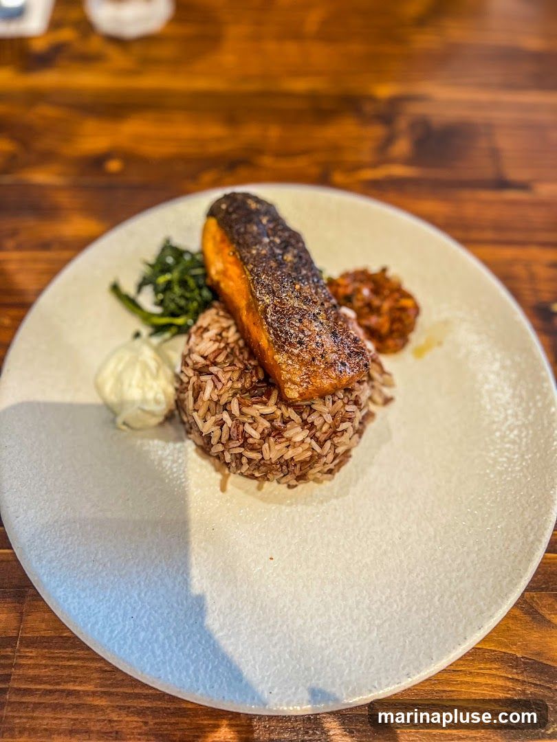 A close-up shot of a plate of food featuring a piece of pan-seared salmon with crispy skin, served atop a mound of brown rice. Accompanying the salmon are some wilted greens, a dollop of white sauce, and a small portion of a chunky relish or salsa. The plate is set on a wooden table.