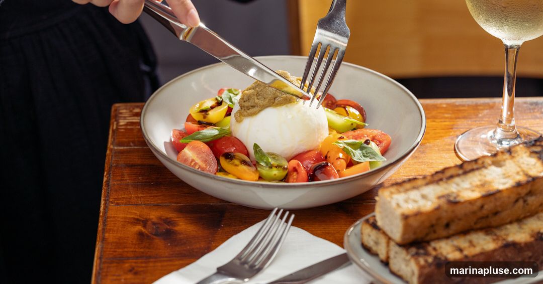 A close-up shot of a burrata cheese dish served with cherry tomatoes and basil, topped with a spread. A glass of white wine and grilled bread are visible on the side. A person is cutting into the burrata with a knife and fork.