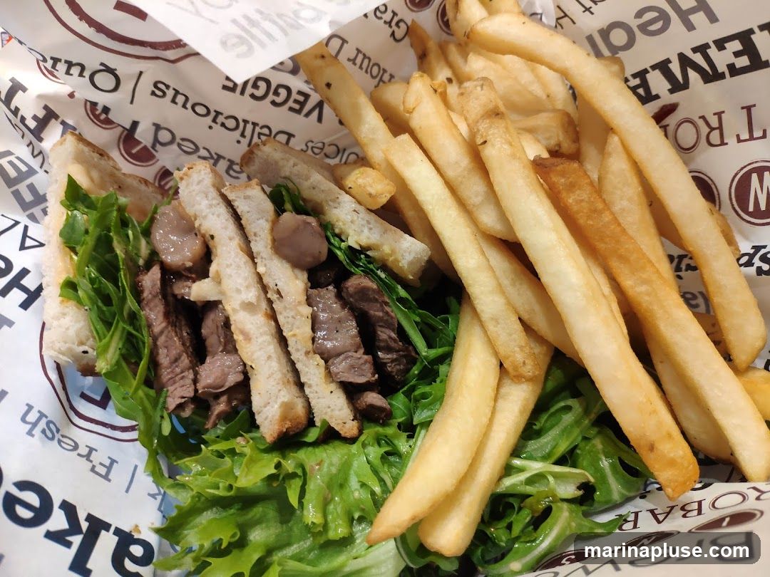 A close-up view of a steak sandwich served with a side of french fries. The sandwich is made with sliced steak, arugula, and a creamy sauce, all on toasted bread. The fries are golden brown and appear to be seasoned. The food is presented on paper with text and logos, suggesting it is from a restaurant or cafe.