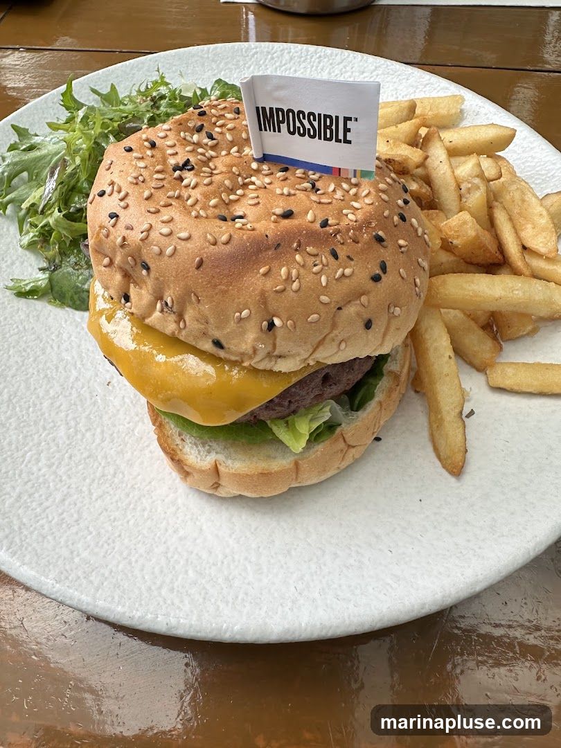 A close-up shot of a plant-based burger, identified by an "Impossible" flag, served on a white plate with a side of french fries and a small salad. The burger has a sesame seed bun, melted cheese, lettuce, and a patty. The fries are golden brown and appear to be thick-cut. The salad consists of mixed greens.