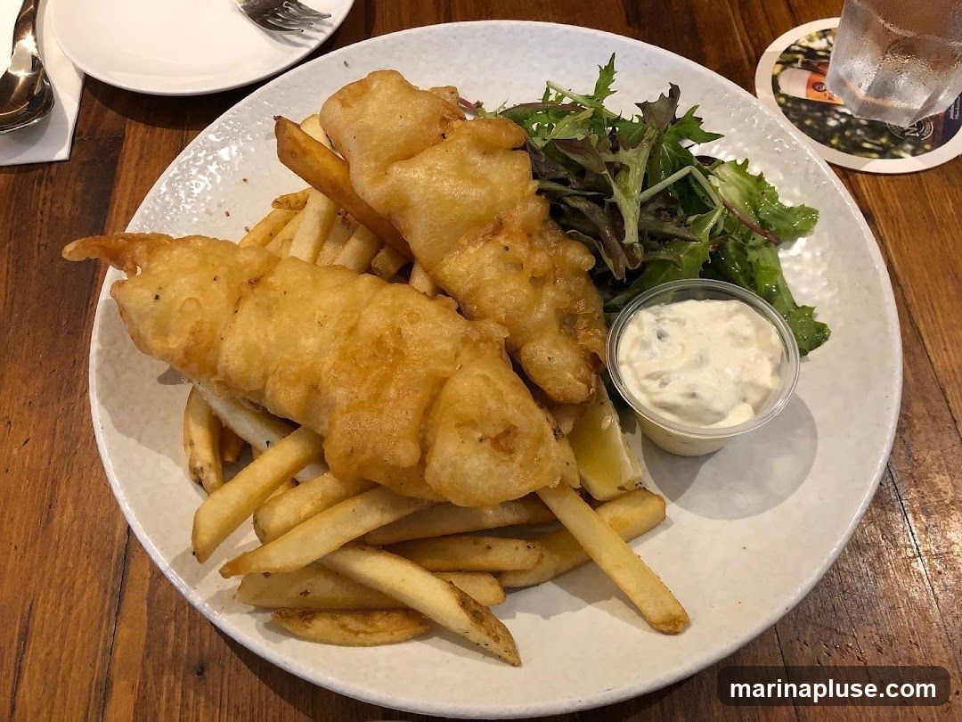 A plate of fish and chips served with a side salad and tartar sauce. The fish is battered and fried, and the chips are thick-cut fries. The dish is presented on a white plate with a textured rim, set on a wooden table. A coaster with a beer advertisement is visible in the background.