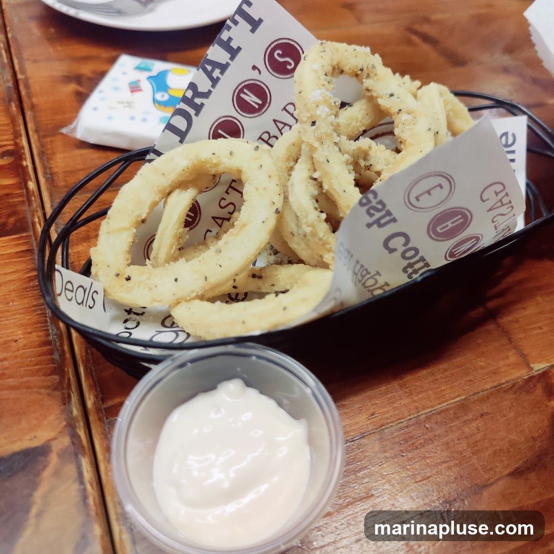A basket of fried calamari rings served with a side of dipping sauce on a wooden table. The calamari is seasoned with herbs and spices. The basket is lined with paper that has a repeating logo and text.