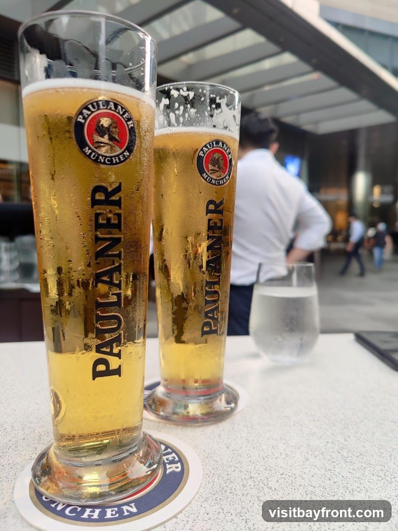 Two tall glasses of Paulaner beer sit on a table, with condensation on the outside of the glasses. In the background, a person in a white shirt is visible, along with other patrons and an outdoor seating area. A glass of water is also on the table.