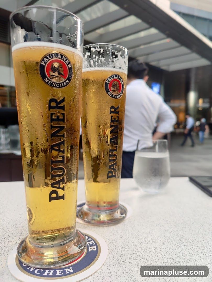 Two tall glasses of Paulaner beer sit on a table, with condensation on the outside of the glasses. In the background, a person in a white shirt is visible, along with other patrons and an outdoor seating area. A glass of water is also on the table.