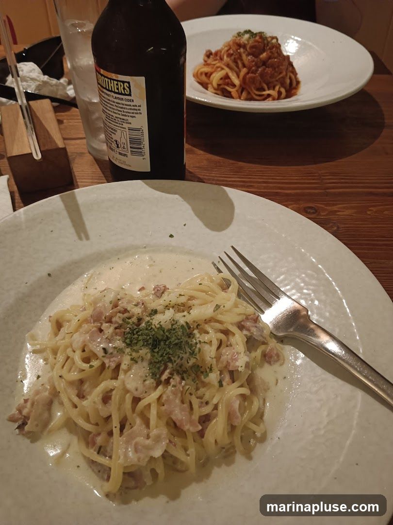 A close-up view of two plates of pasta dishes and a bottle of cider on a wooden table. The plate in the foreground features spaghetti carbonara, garnished with parsley. The plate in the background contains a pasta dish with a red sauce. A bottle of cider with a label indicating it is from "Brothers" is also visible.