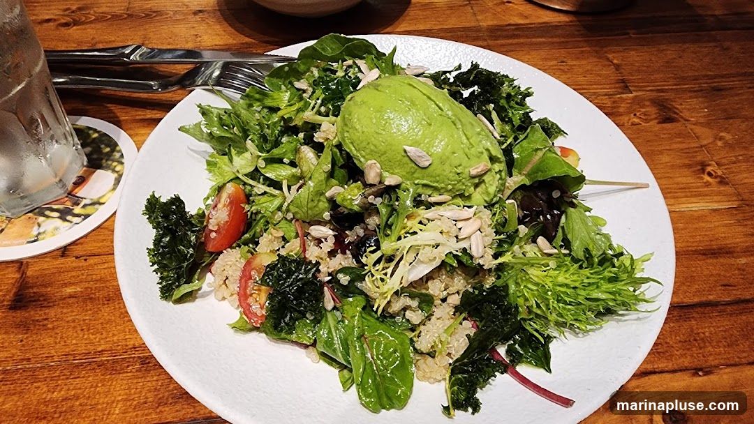 A close-up shot of a vibrant salad served on a white plate. The salad features a generous portion of avocado, quinoa, cherry tomatoes, kale, and mixed greens, sprinkled with sunflower seeds. The plate is set on a rustic wooden table, with a glass of water and cutlery visible in the background.