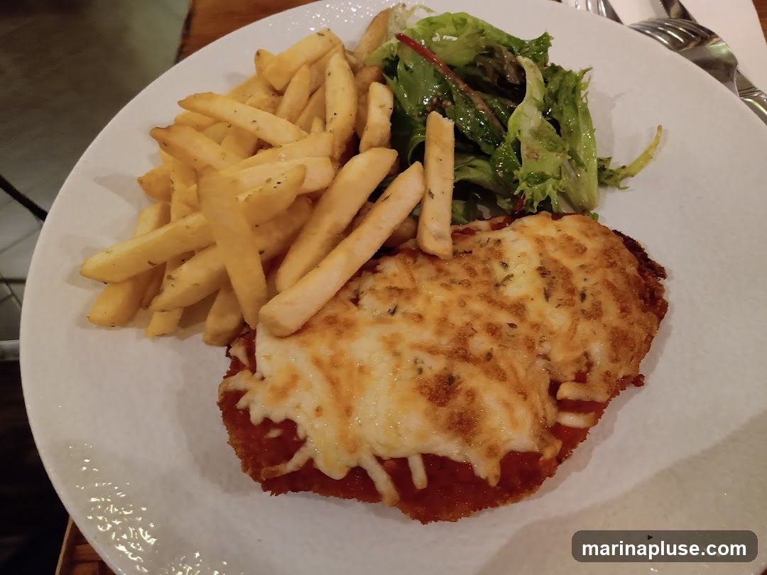 A close-up shot of a plate of food featuring chicken parmesan, a side of french fries, and a small green salad. The chicken is breaded and topped with melted cheese and tomato sauce. The fries are golden brown and seasoned. The salad consists of mixed greens with a light dressing.