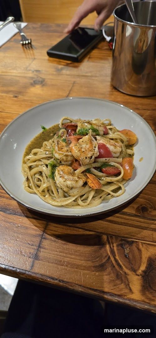 A plate of linguine pasta with shrimp and cherry tomatoes, tossed in a green sauce. The dish is served on a wooden table, with a fork and a smartphone visible in the background.