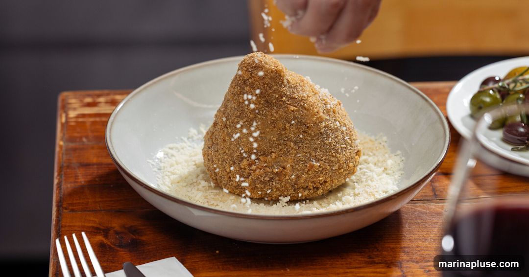 A close-up shot shows a hand sprinkling grated cheese over a large, breaded arancini ball in a bowl. The arancini is coated in breadcrumbs and appears to be resting on a bed of more grated cheese. To the right, a small dish contains olives, and a glass of red wine is visible in the foreground. The food is placed on a rustic wooden table.