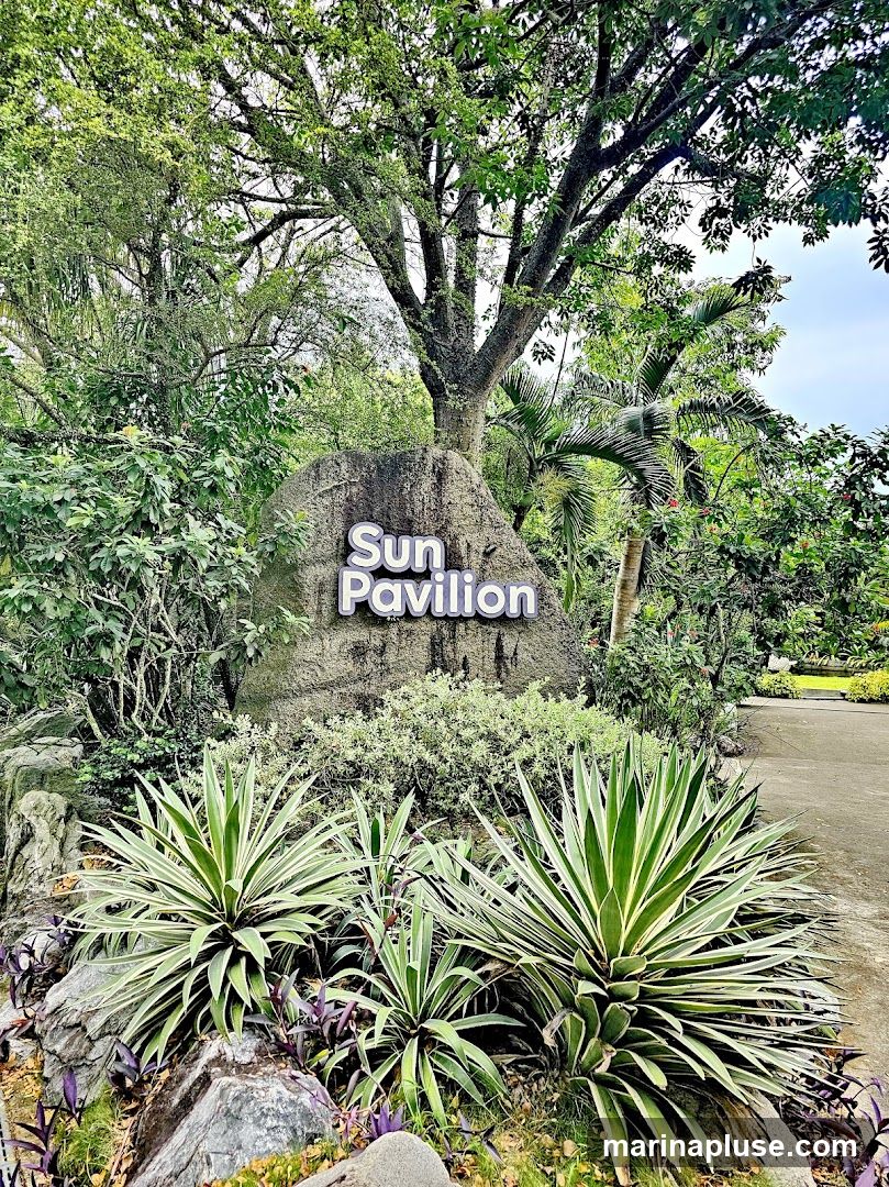 A large rock with the words "Sun Pavilion" in raised lettering is surrounded by lush greenery and tropical plants. The sign is set in a garden-like environment with a paved path visible to the right.