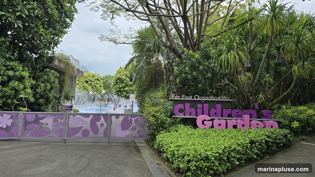 A sign reads "Far East Organization Children's Garden" in large purple and pink letters, set against a backdrop of lush green foliage. In the background, a water play area with fountains and children can be seen. A decorative purple gate with floral and abstract patterns is in the foreground.