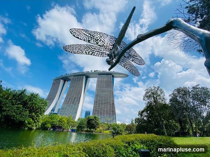 A large, intricate metal sculpture of a dragonfly is prominently displayed in the foreground, with its wings spread wide. Behind the sculpture, the iconic Marina Bay Sands hotel complex in Singapore is visible, featuring its distinctive three towers topped by a ship-like structure. The scene is set against a bright blue sky with scattered white clouds. Lush green trees and a body of water are in the mid-ground, suggesting a park or garden setting.