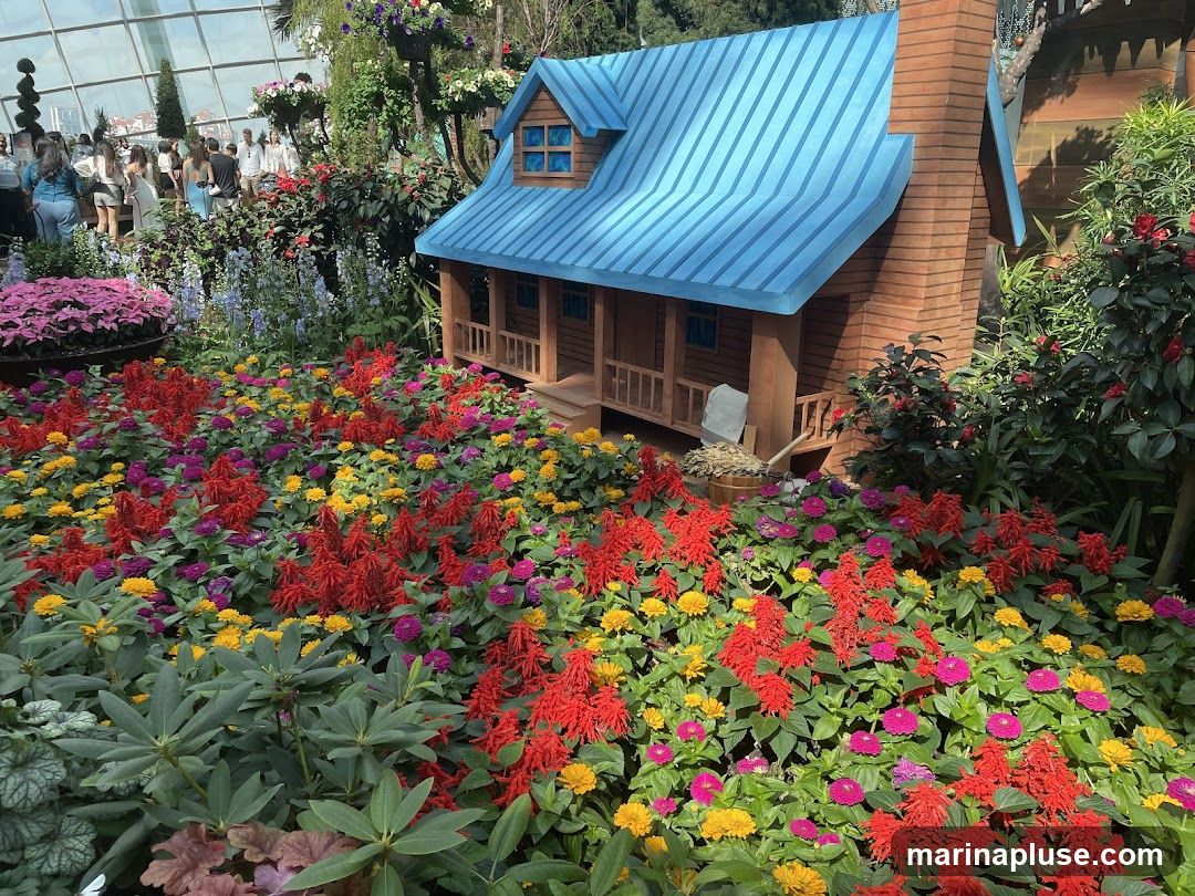 A miniature wooden house with a blue roof and brick chimney is surrounded by a vibrant display of flowers. The flowers are a mix of red, yellow, and purple, with lush green foliage. In the background, people are visible walking through a large greenhouse or botanical garden.