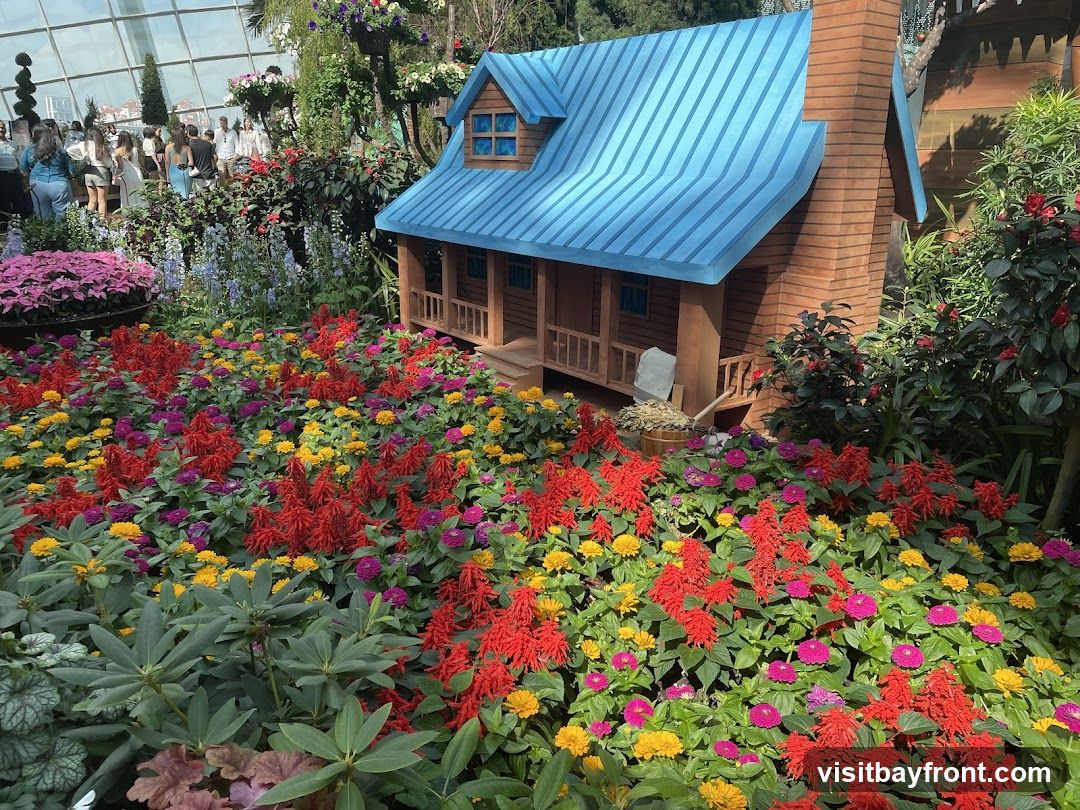 A miniature wooden house with a blue roof and brick chimney is surrounded by a vibrant display of flowers. The flowers are a mix of red, yellow, and purple, with lush green foliage. In the background, people are visible walking through a large greenhouse or botanical garden.