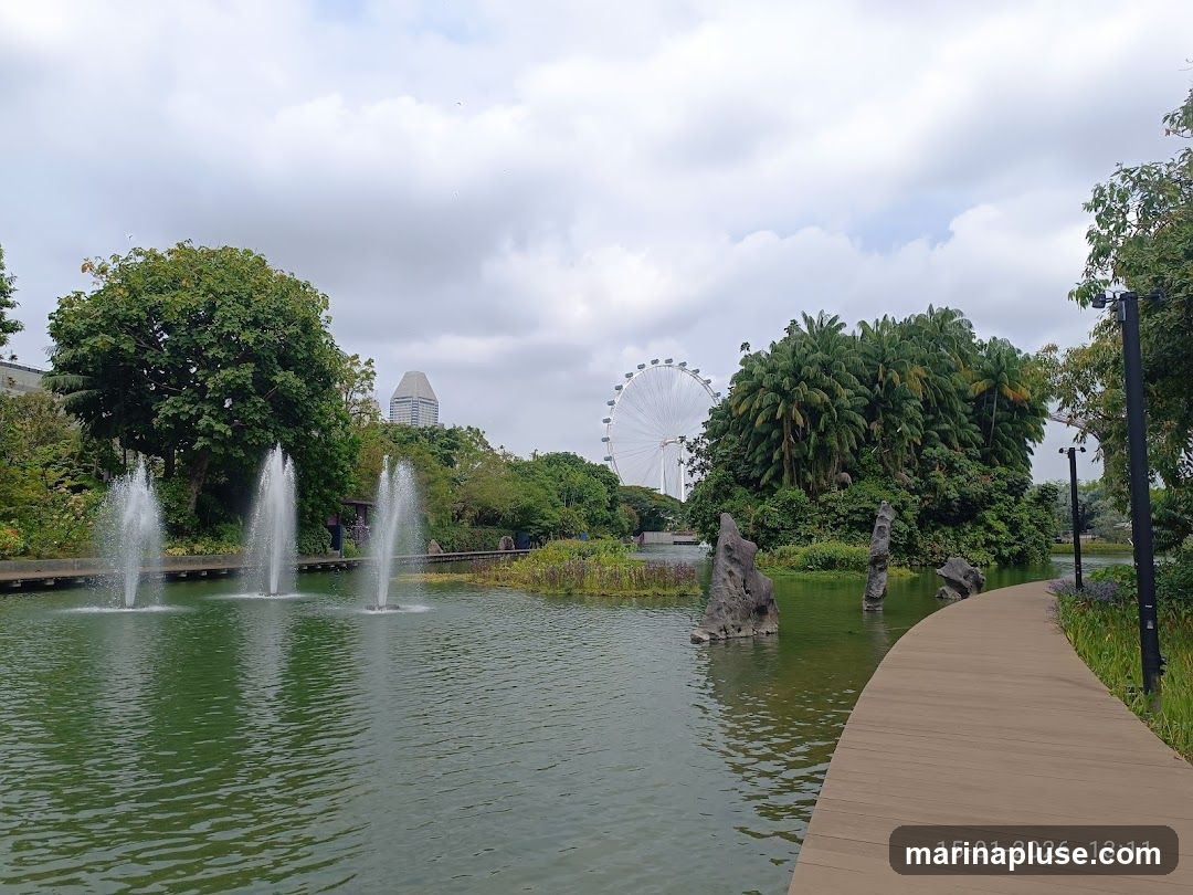 A serene park scene featuring a tranquil body of water with three active fountains. A wooden boardwalk curves along the right side of the water. In the background, lush greenery, palm trees, and a large Ferris wheel are visible under a cloudy sky. A modern building can be seen in the distance. The date stamp '15-01-2026 13:11' is present in the lower right corner.