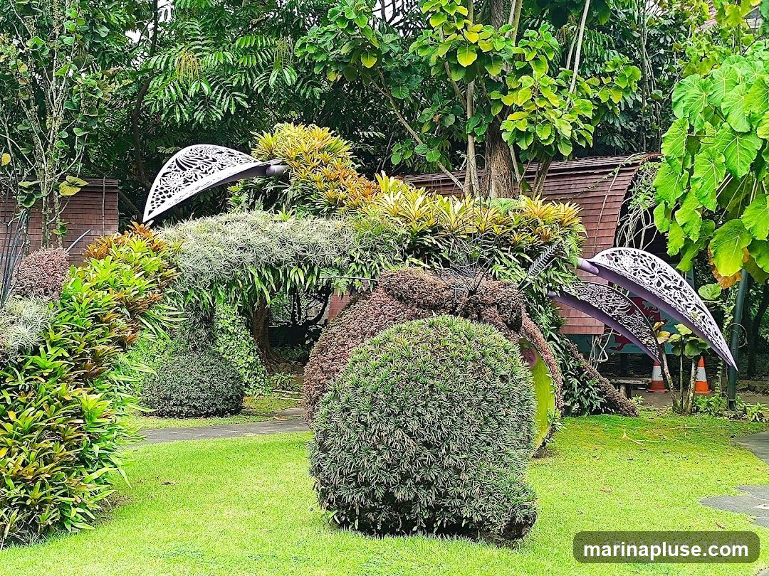 A large, intricately shaped topiary sculpture resembling a bird or insect is the centerpiece of a lush garden. The sculpture is adorned with various plants, including air plants and small shrubs, and features decorative metal wings. Behind the sculpture, a wooden structure with a curved roof is visible, surrounded by dense foliage and trees. The ground is covered with neatly manicured green grass, and a pathway winds through the garden. The scene is bathed in natural light, highlighting the vibrant greens of the plants.