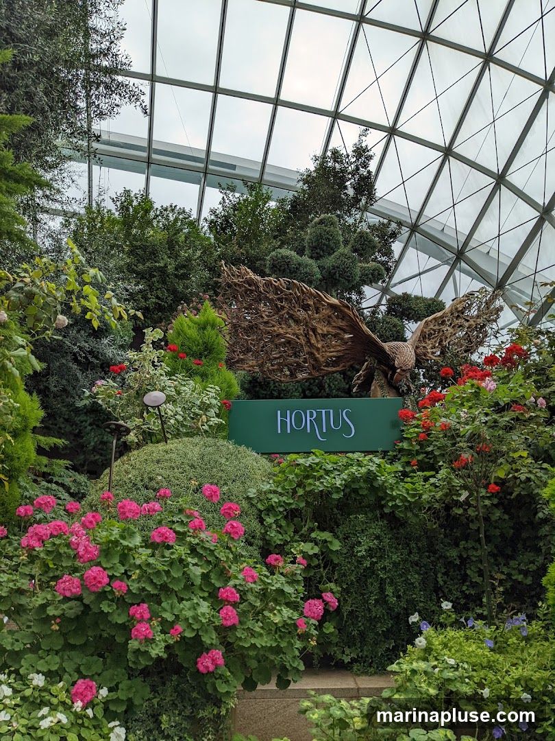 A lush garden display featuring a large metal sculpture of a bird with outstretched wings. The sculpture is positioned behind a green sign that reads "HORTUS" in white lettering. The garden is filled with a variety of plants, including vibrant pink geraniums, red roses, and various green shrubs and trees, all under a geodesic glass roof.