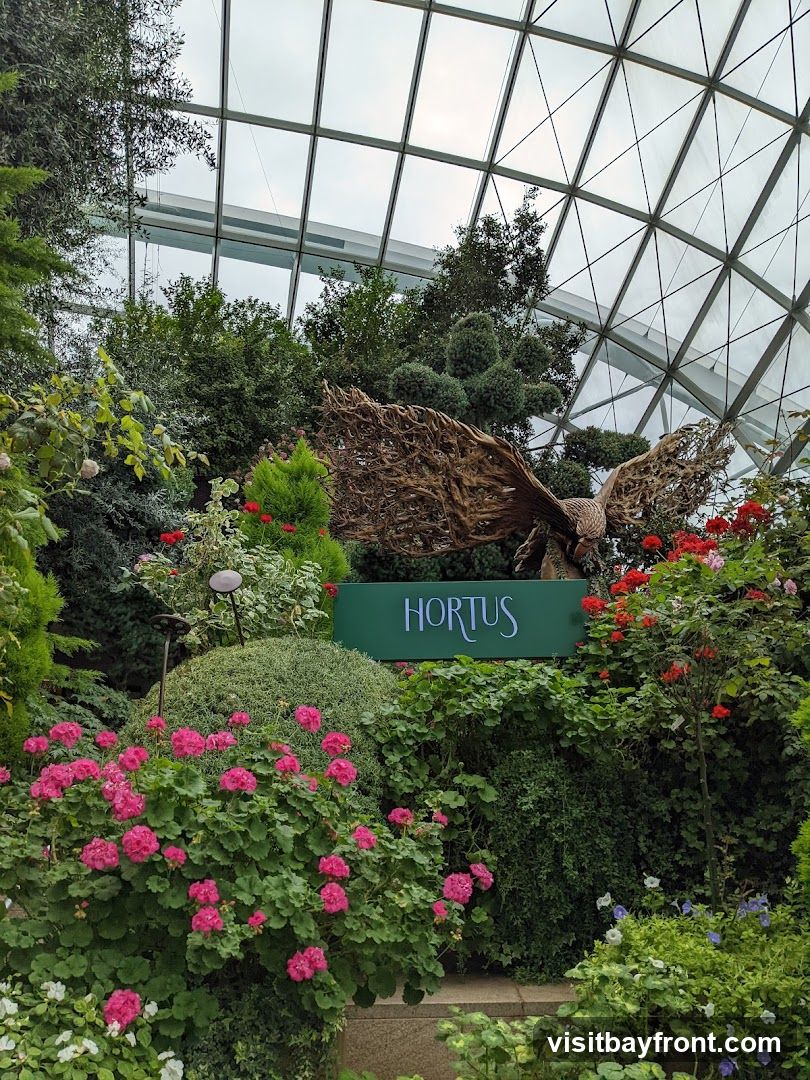 A lush garden display featuring a large metal sculpture of a bird with outstretched wings. The sculpture is positioned behind a green sign that reads "HORTUS" in white lettering. The garden is filled with a variety of plants, including vibrant pink geraniums, red roses, and various green shrubs and trees, all under a geodesic glass roof.