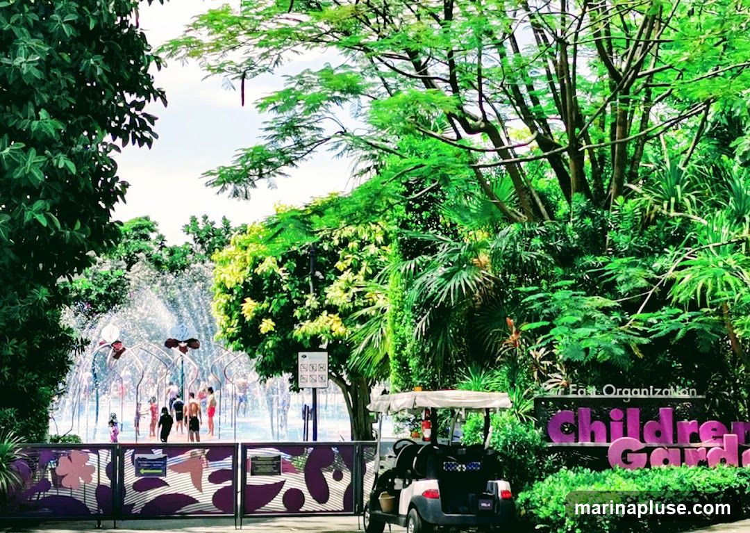 A view of the entrance to a Children's Garden with a water play area in the background. Children are seen playing in the water fountains. A golf cart is parked near a sign that reads "East Organization Children's Garden". Lush greenery surrounds the area.