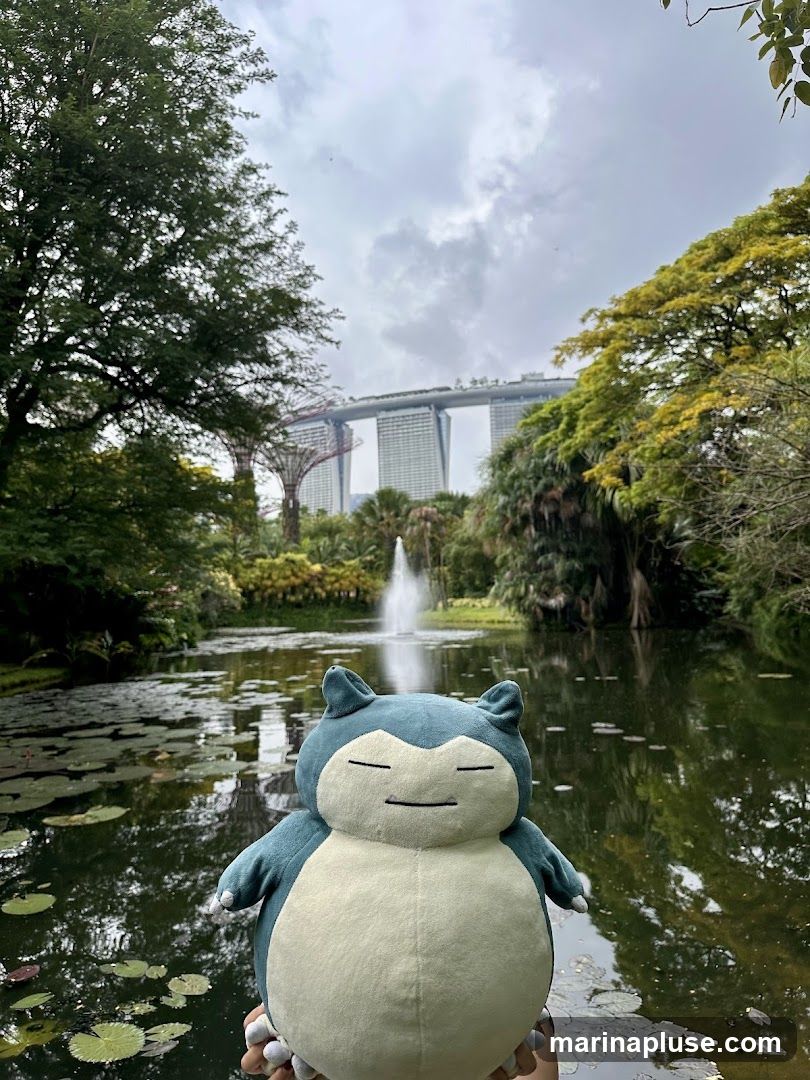 A Snorlax plush toy is held in the foreground, with a scenic view of Gardens by the Bay in Singapore in the background. The Gardens feature lush greenery, a tranquil pond with lily pads, a fountain, and the iconic Marina Bay Sands hotel complex visible in the distance under a cloudy sky.