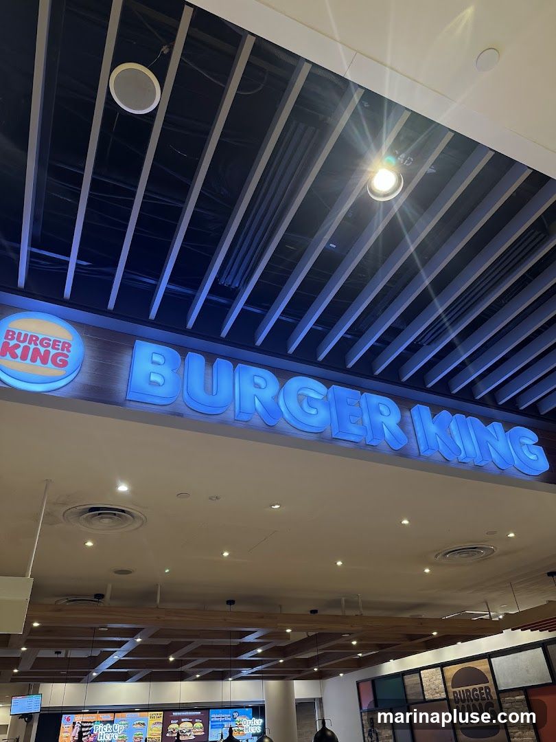 A Burger King sign with blue illuminated lettering is displayed prominently. The iconic Burger King logo is visible on the left side of the sign. The ceiling above features exposed beams and lighting fixtures. Below the main sign, a menu board with various food items and prices is partially visible.