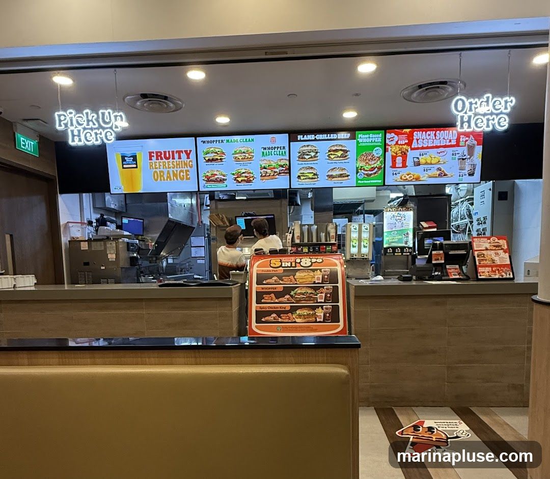 A view of a fast-food restaurant counter with digital menu boards displaying various burgers and drinks. Signs indicate "Pick Up Here" and "Order Here". Two employees are visible behind the counter. A promotional stand with meal deals is also present.