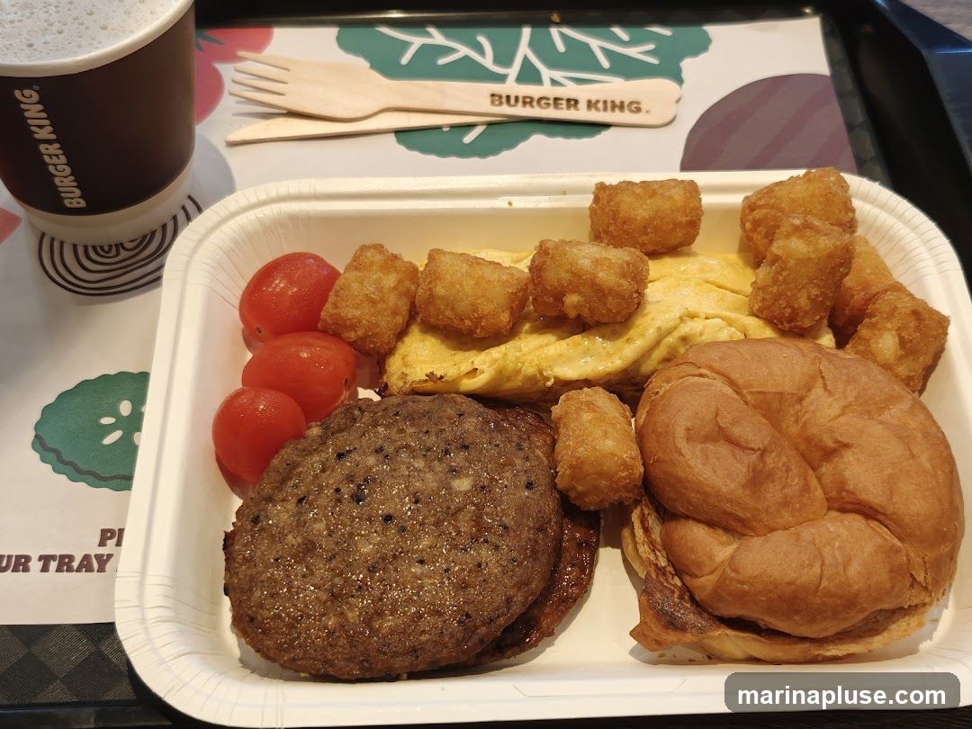 A Burger King breakfast meal is presented on a white tray. The meal includes a sausage patty, scrambled eggs, cherry tomatoes, tater tots, and a croissant sandwich. A cup with the Burger King logo is visible on the left, and wooden cutlery is placed on a tray liner with the Burger King logo.