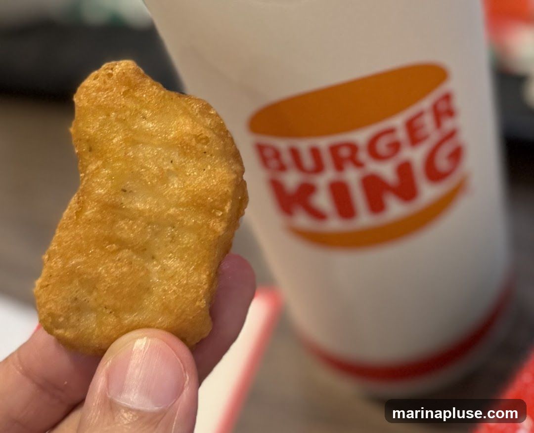 A close-up shot of a single golden-brown chicken nugget held between fingers, with a Burger King branded cup blurred in the background. The cup features the iconic orange and red Burger King logo.
