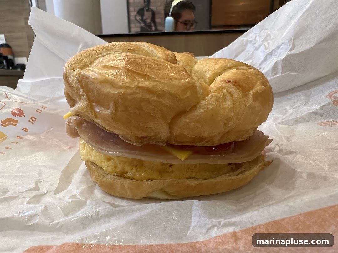 A close-up shot of a breakfast sandwich on a croissant, wrapped in white paper. The sandwich contains a folded omelet, slices of ham, and a slice of cheese. The paper wrapping has an orange logo with a stylized chicken and text that is partially visible. In the background, a person is reflected in a mirror or window, and there is a framed picture on the wall.