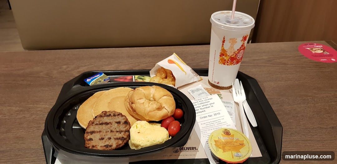A Burger King breakfast meal is presented on a black tray. The meal includes pancakes, a sausage patty, scrambled eggs, cherry tomatoes, a croissant, a side of tater tots, a cup with a straw, and a small container of maple syrup. A receipt and cutlery are also on the tray.