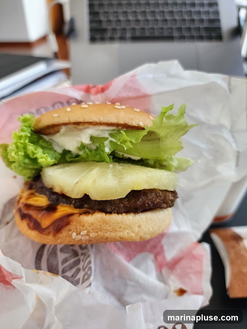 A close-up shot of a burger with lettuce, pineapple, cheese, and a patty, wrapped in branded paper. A laptop and papers are visible in the blurred background.